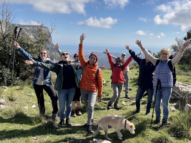Wandergruppe mit erhobenen Armen auf einer Bergwiese, dazu ein Hund, dahinter Felsen und das Meer