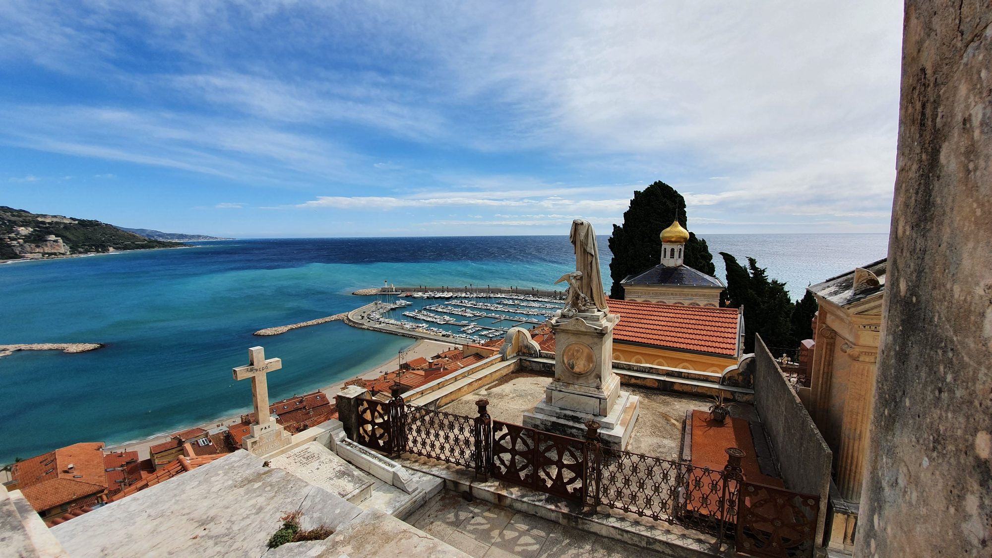 Vue plongeante sur le port et la baie turquoise, au premier plan une statue de pierre et une croix