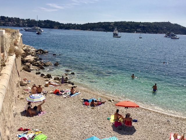 Kiesstrand in einer Bucht mit Sonnenschirmen, Badeg&auml;sten und ankernden Booten