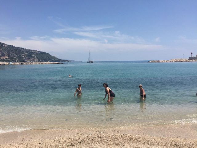 Drei Menschen stehen im flachen Wasser vor einem Kiesstrand