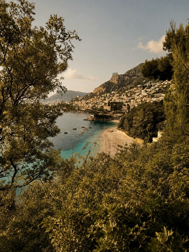 T&uuml;rkise Bucht mit Kiesstrand und H&uuml;gelort an der C&ocirc;te d&rsquo;Azur, durch mediterrane B&auml;ume gesehen
